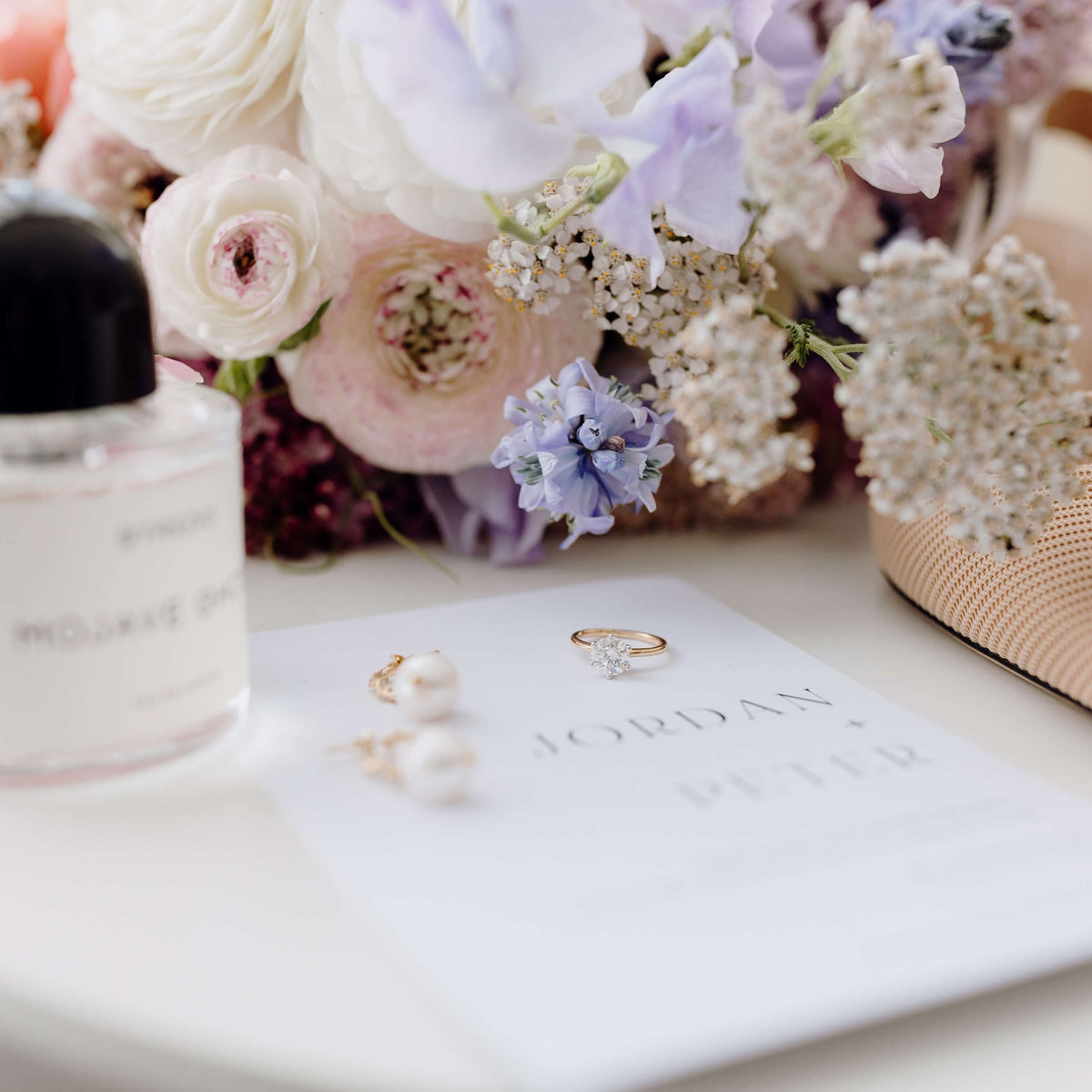 Engagement ring and pearl earrings resting on wedding invitation with bouquet behind