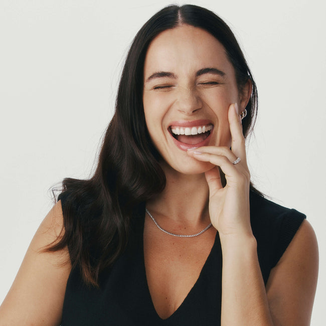 Woman laughing wearing diamond necklace, earrings and ring against a neutral background
